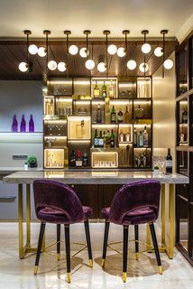 two purple chairs sitting in front of a bar with bottles on the shelves and lights above it