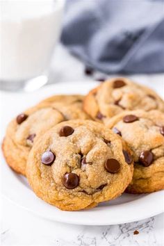 three chocolate chip cookies on a white plate with a glass of milk in the background