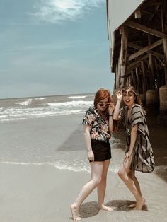 two young women walking on the beach next to a pier