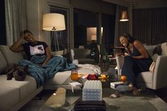 two women sitting on couches in a living room with food and drinks scattered around