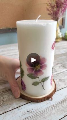 a hand holding a white candle with purple flowers on it and a wooden slice next to it