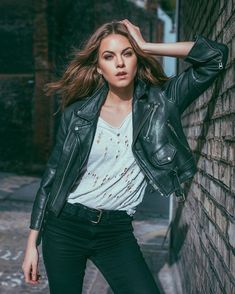 a woman leaning against a brick wall wearing a black leather jacket and white t - shirt