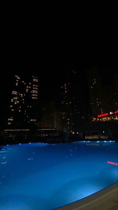 an empty swimming pool in the middle of a city at night with skyscrapers lit up behind it