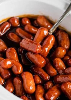 hot dogs are being cooked in a white bowl with sauce on the side and a spoon sticking into them