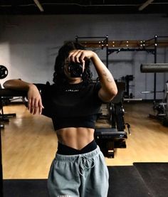 a woman taking a selfie with her camera in a gym area, wearing sweatpants and crop tops