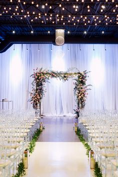 an indoor wedding ceremony setup with white chairs and floral arrangements on the back drop cloth drapes