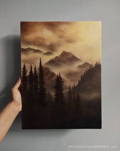 a person holding up a painting with mountains and trees in the background on a gray wall