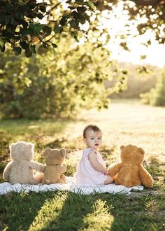 a baby sitting in the grass with three teddy bears next to her on a blanket