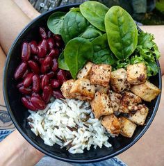 a person holding a black bowl filled with rice, beans and greens next to other foods