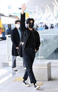 a man in black jacket and sunglasses walking on sidewalk with his hand up to the air