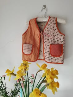 two vests hanging on the wall with yellow flowers in front of them and a vase full of daffodils