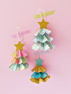 two paper christmas trees hanging from strings on a pink background with gold and silver stars