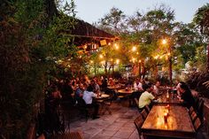 people sitting at tables in an outdoor restaurant with lights strung from the ceiling and greenery