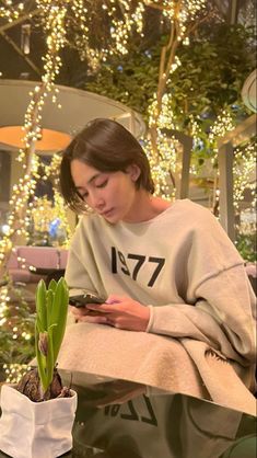 a woman sitting down looking at her cell phone next to a potted plant with lights in the background