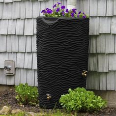 a tall black planter with purple flowers in it sitting next to a building and grass