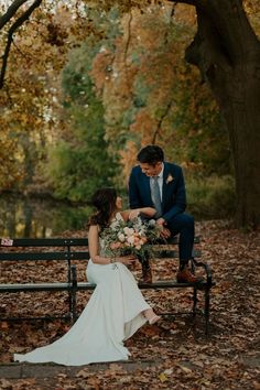 a bride and groom sitting on a bench in the park with leaves all around them