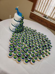 a peacock cake and cupcakes on a white tablecloth with windows in the background