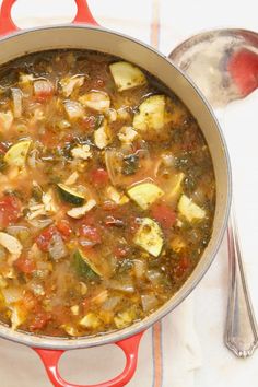 a pot filled with vegetable soup on top of a white table cloth next to a spoon