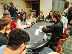 a group of people sitting around a black table playing cards in a room full of other people