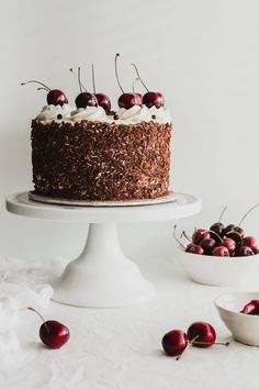 a chocolate cake with cherries on top sitting on a white pedestal next to bowls of cherries