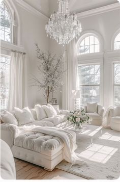 a living room with white furniture and chandelier hanging from the ceiling in front of two large windows