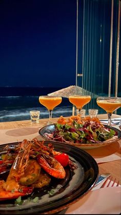 two plates of food on a table with wine glasses and an ocean view in the background