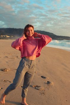 a woman standing on top of a sandy beach next to the ocean with her hands behind her head