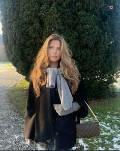 a woman standing in front of a tree holding a handbag and wearing a scarf