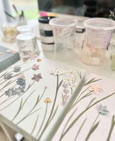 an open book sitting on top of a table next to some paint bottles and flowers