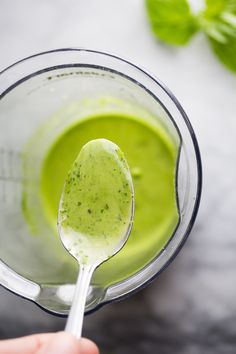 a hand holding a spoon in a blender filled with green liquid and basil leaves