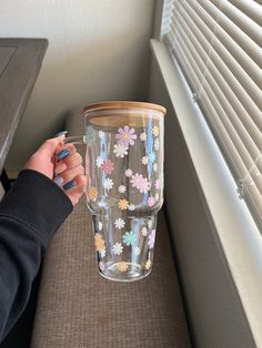 a person holding up a glass cup with flowers painted on the bottom and sides, in front of a window