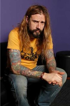 a man with long hair and tattoos sitting on a couch in front of a purple wall