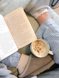 a cup of coffee sitting next to an open book on top of a bed with someone's feet propped up