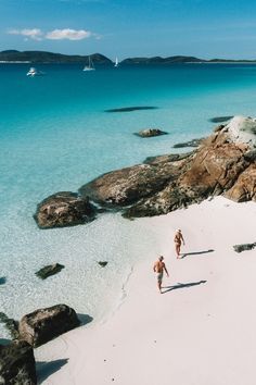 two people are walking on the beach in front of some rocks and clear blue water