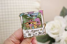 a person holding up a pin with the words the magic house on it and flowers in the background