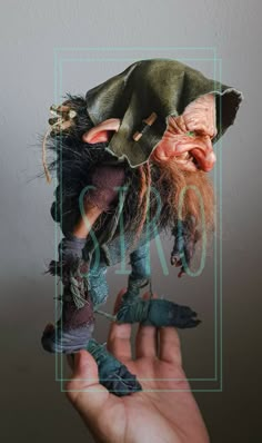 a hand holding an old troll doll with green hair and red beard, in front of a white wall