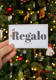 a person holding up a card with the word regalo in front of a christmas tree