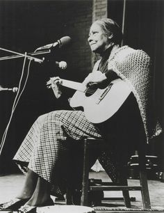 a black and white photo of a woman sitting in front of a microphone playing an acoustic guitar