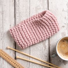 a pink knitted hat next to two knitting needles and a cup of coffee on a white wooden table