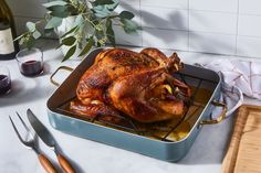 a roasted turkey in a roasting pan on a counter next to wine glasses and utensils