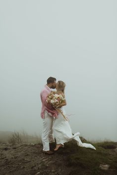 a bride and groom kissing on top of a hill in the foggy weather with their bouquet