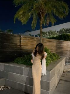 a woman in a long dress standing next to a wall and palm tree at night