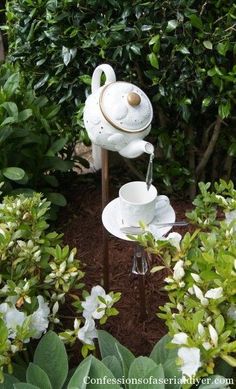 a white tea pot and cup on a stand in the middle of some bushes with flowers around it