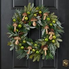 a wreath with green and gold ornaments hanging on a black front door, surrounded by greenery