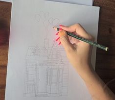 a woman is drawing a house with colored pencils on top of the paper and holding a pen in her hand