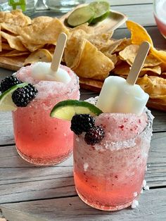 two glasses filled with fruit and ice sitting on top of a table next to chips