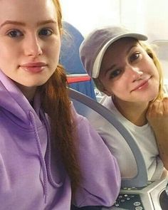 two young women sitting next to each other on a plane, one is wearing a hat and the other wears a purple sweatshirt