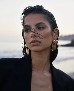 a woman with long hair wearing gold earrings and a black jacket on the beach at sunset