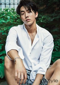 a young man sitting on the ground with his hands in his pockets, wearing a white shirt and patterned shorts