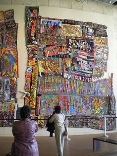 two women standing in front of a large wall covered with colorful fabric art pieces,